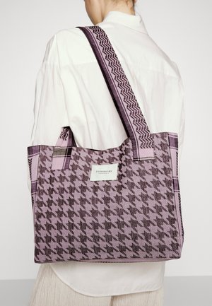 Persona con camisa blanca lleva un bolso tipo tote grande con estampado de pata de gallo en morado y negro, con una correa ancha y una etiqueta blanca.