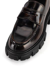 Brązowy mokasyn ze skóry o błyszczącym wykończeniu, z przeszywanym wzorem, grubą gumową podeszwą oraz wyraźnym paskiem na cholewce.