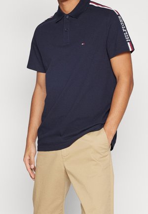 Mann trägt dunkelblaues Poloshirt mit rot-weiß-blauem Streifen und "Hilfiger" am Ärmel, kombiniert mit beigen Hosen, Hand in der Tasche.