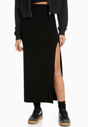 Femme portant une jupe maxi noire avec une fente haute sur le côté, des chaussettes noires, des baskets noires et grises, et une veste courte noire.