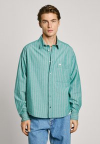 Camicia button-up a righe verdi con vestibilità rilassata, caratterizzata da righe orizzontali bianche e rosse, e una tasca sul petto. Indossata con jeans blu.
