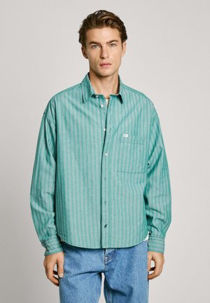 Green striped button-up shirt with a relaxed fit, featuring horizontal stripes in white and red, and a chest pocket. Worn with blue jeans.