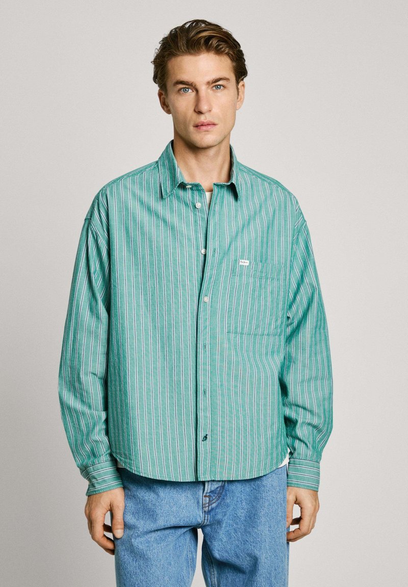 Camicia button-up a righe verdi con vestibilità rilassata, caratterizzata da righe orizzontali bianche e rosse, e una tasca sul petto. Indossata con jeans blu.