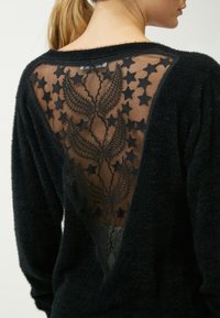 Maglione nero morbido con schiena trasparente decorata con stelle ricamate e motivi floreali. Design a V con dettagli in tessuto testurizzato.