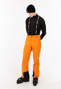Man in een zwarte beanie, zwart langemouwen shirt met bretels, feloranje skibroek en zwarte skischoenen tegen een witte achtergrond.