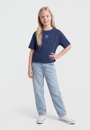 Blond pige står med hænderne på hoften iført navyblå Converse T-shirt, lyseblå jeans og hvide sneakers mod hvid baggrund.