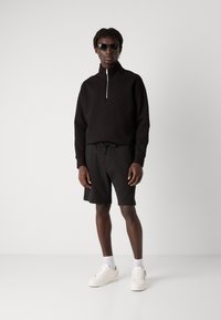 Felpa nera con zip e colletto alto, abbinata a pantaloni corti neri texture e sneaker bianche. Gli accessori includono occhiali da sole scuri.