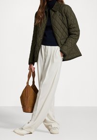 Persoon in een olijfkleurige gewatteerde jas, marineblauwe trui, brede witte broek en witte sneakers, met een bruin leren handtas in de hand en de andere hand in de zak.