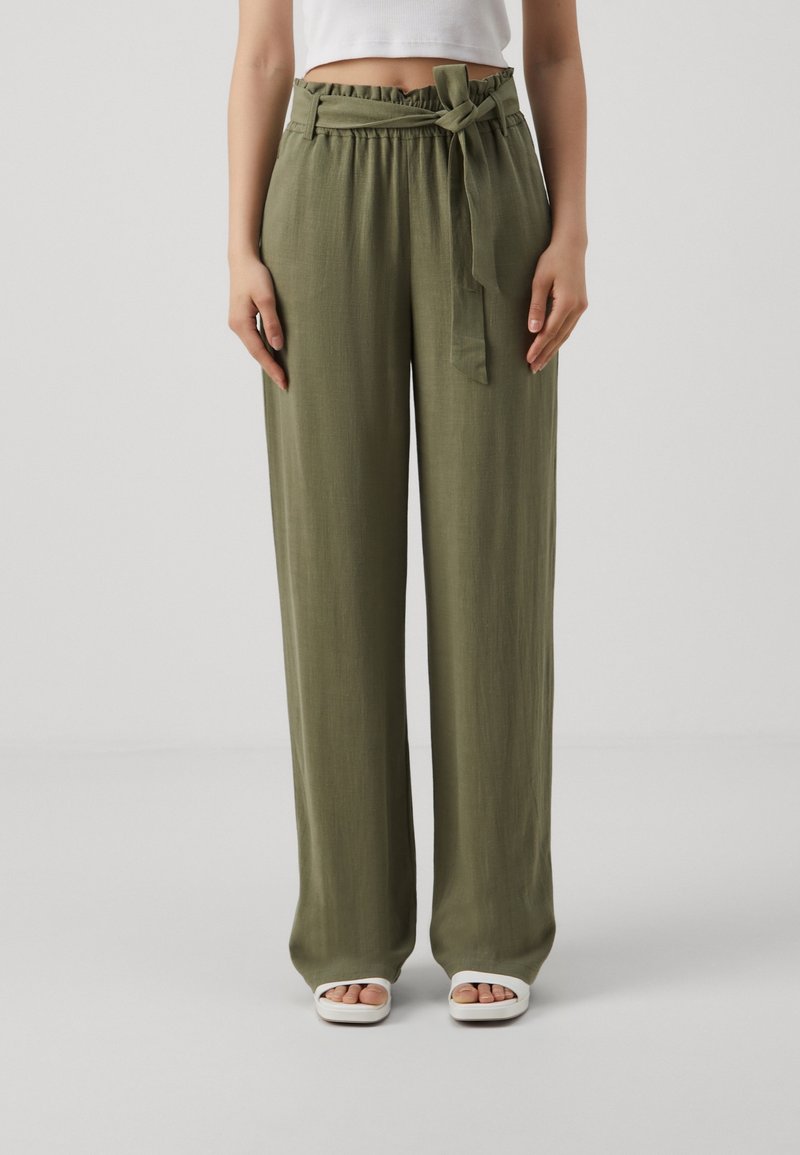 Pantalon vert, léger, à jambes larges avec une taille cintrée et un détail noué. Le tissu a une texture lisse. Associé à des chaussures blanches à bouts ouverts.