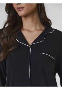 Svart pyjamasöverdel med vit piping, knappstängning framtill och bröstficka, med ribbad textur och klassisk kragsdesign.