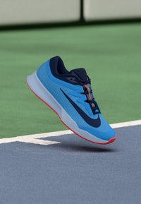 Blå sportskor med en strukturerad ovandel, med en mörkblå swoosh-detalj och orange sula. Positionerade ovanför en grå plan yta.