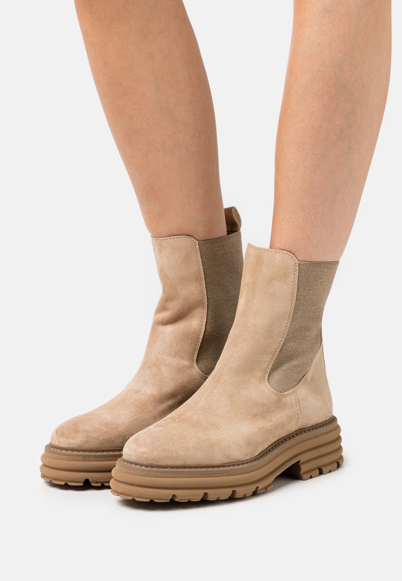 Bottines beiges en daim avec semelles épaisses à rainures portées sur jambes nues, avec des panneaux latéraux élastiques et des languettes à l’arrière.