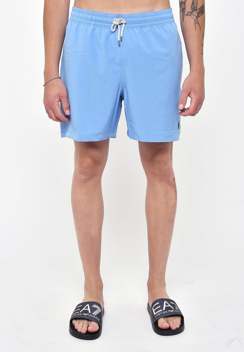 Pantaloni da bagno azzurri chiari con cintura elastica e orlo corto. Indossati con sandali neri a infradito con testo del logo bianco. Texture del tessuto liscia.