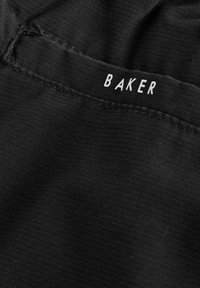 Nahaufnahme von schwarzem Stoff mit sichtbaren Nähten und einem kleinen weißen Etikett mit der Aufschrift "BAKER" an einer gefalteten Naht.