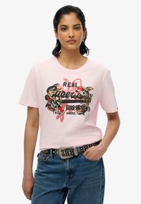Camiseta de algodón de color rosa claro con cuello redondo, que presenta un diseño gráfico de coloridos peces koi y texto. Combinada con unos vaqueros de mezclilla azules y un cinturón negro.