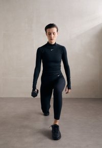 Schwarzes Sportoberteil mit langen Ärmeln und gerippten Leggings mit Nike-Logo; hält eine sechseckige Hantel; schwarze Turnschuhe; minimalistischer Hintergrund.