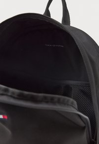 Črn platnen nahrbtnik z večjim glavnim predelom, mrežnim žepom in logotipom blagovne znamke "TOMMY HILFIGER" na notranji oblogi.