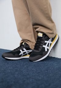 Black and gray athletic shoes with white stripes and yellow accents, featuring a mesh upper and rubber sole, placed on a blue carpet.