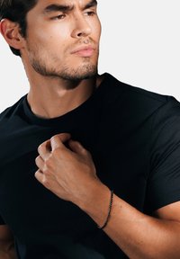 Musta lyhythihaisuus t-paita, jossa on sileä tekstuuri; käsi puristaa kangasta ja paljastaa mustan helmikintaan ranteessa.