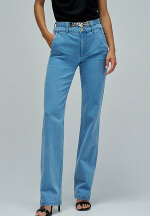 Femme portant un jean bleu clair taille haute, coupe droite, un top noir sans manches, des sandales noires à talons et une ceinture noire avec une boucle métallique.