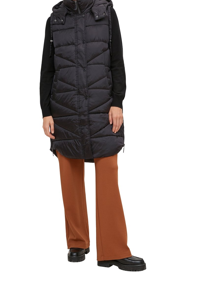 Personne portant une longue veste sans manches noire matelassée par-dessus des manches longues noires, un pantalon marron à jambes larges et des chaussures noires à semelle épaisse.
