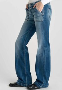Jean bleu à jambes larges avec une finition décolorée, cinq poches, bouton en métal et détails de couture. Associés à des ballerines noires en cuir verni.