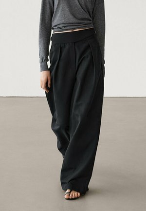 Czarne spodnie o szerokich nogawkach z plisami i wysokim stanem, zestawione z dopasowaną szarą bluzką z długim rękawem, na neutralnym tle.