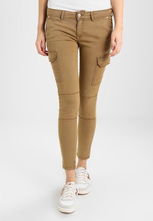 Frau trägt eine schmal geschnittene hellbraune Cargohose mit Klapptaschen und weiße Sneakers, geht vor einem grauen Hintergrund.