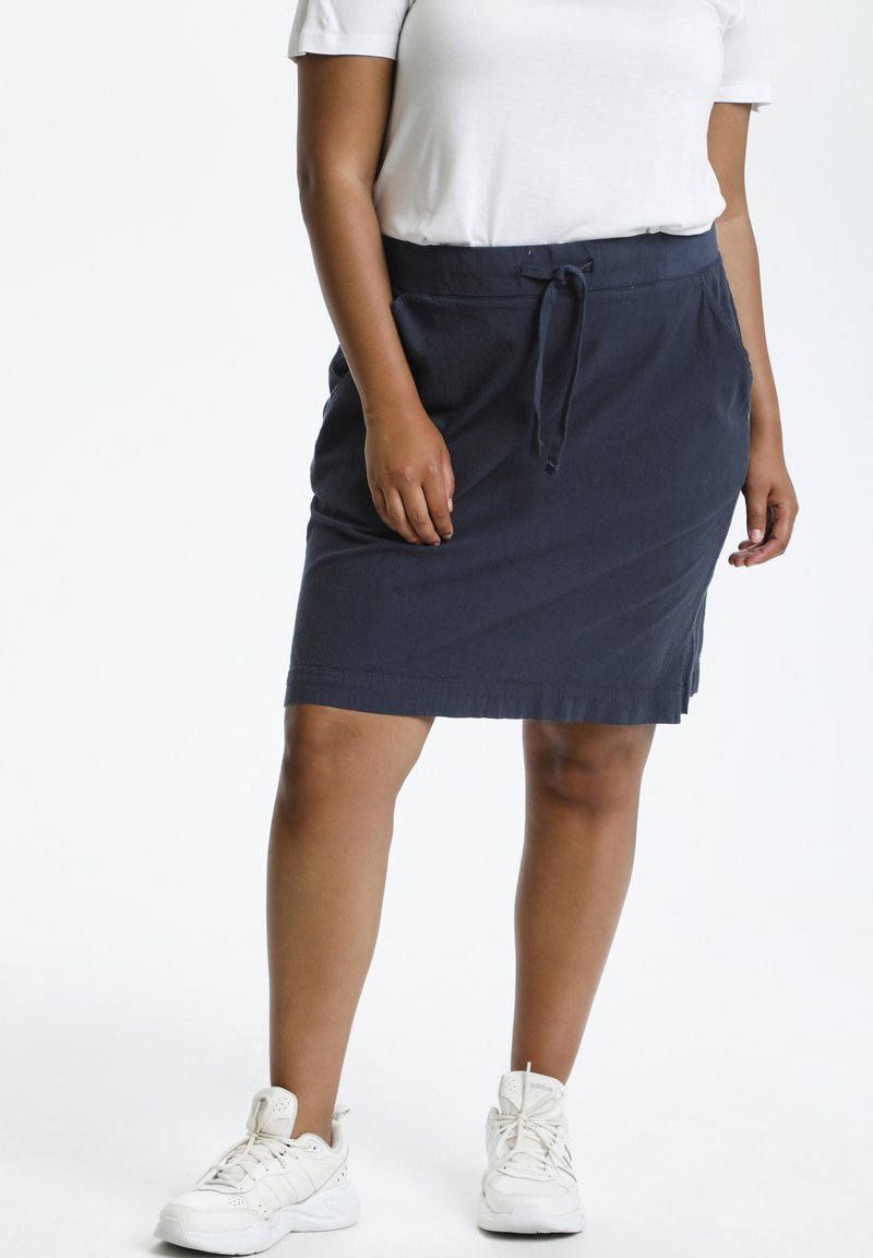 Jupe en coton bleu marine avec une taille à cordon de serrage, des poches latérales et une coupe droite, portée avec des baskets de sport blanches.