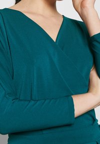 Teal inslagstopp med V-ringning, långärmad och mjukt texturerat tyg, med rynkade detaljer på axlarna.