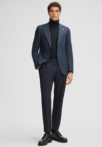 Man in een donkerblauwe geblokte blazer, zwarte coltrui, slanke marineblauwe broek en zwarte leren veterschoenen staand tegen een effen achtergrond.