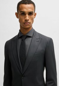 Completo nero su misura con una vestibilità strutturata, caratterizzato da rever a punta e tessuto texture. Accompagnato da una cravatta nera uniforme e una camicia a righe.