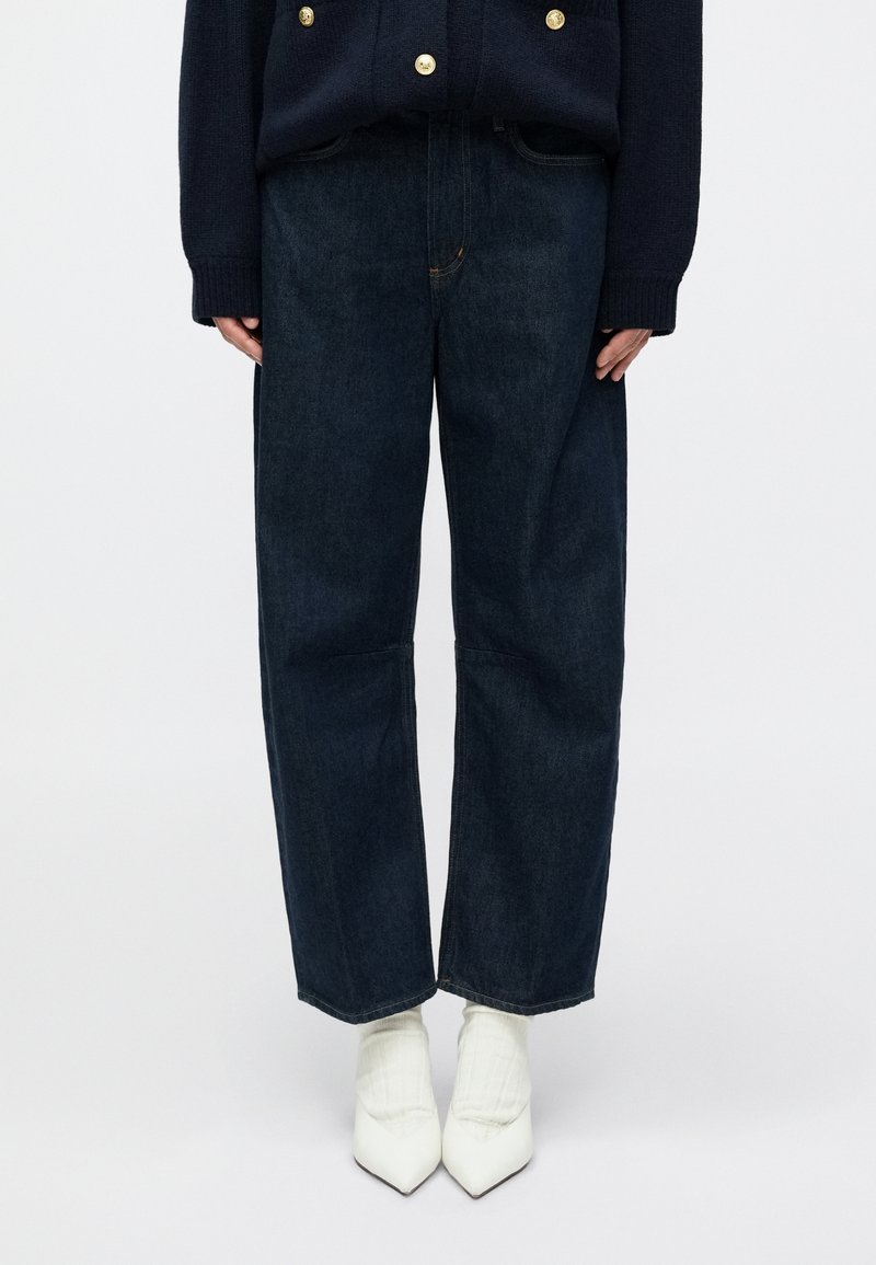 Sötétkék, laza szabású farmernadrág egyenes szárakkal, finom varrási részletekkel, fehér hegyes orrú cipőkkel és bordázott zoknikkal párosítva.