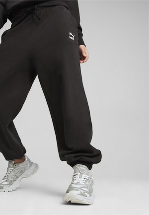 Pantalones deportivos negros con cinturilla elástica, bolsillos laterales y un logo blanco de Puma en el muslo. Llevados con zapatillas deportivas plateadas.