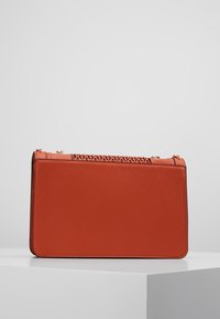 Prostokątna torebka z pomarańczowej skóry o gładkim wykończeniu. Charakteryzuje się misternym wyciętym wzorem na górze oraz złotymi elementami ozdobnymi.