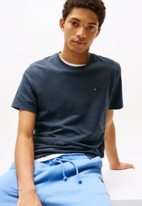 Ung man som sitter, klädd i en mörkblå kortärmad t-shirt och ljusblå mjukisbyxor med dragsko, mot en enkel vit bakgrund.