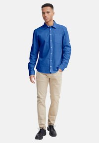 Blaues langärmliges Hemd mit Knöpfen und Brusttasche, kombiniert mit beigen Chinos und schwarzen Schnürschuhen. Glatte Stoffoberfläche.