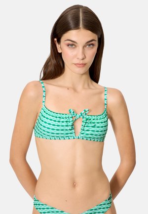 Joven con cabello largo y castaño, vistiendo un bikini de top con patrón ondulado verde y blanco, con tirantes delgados y nudo frontal, sobre un fondo liso.