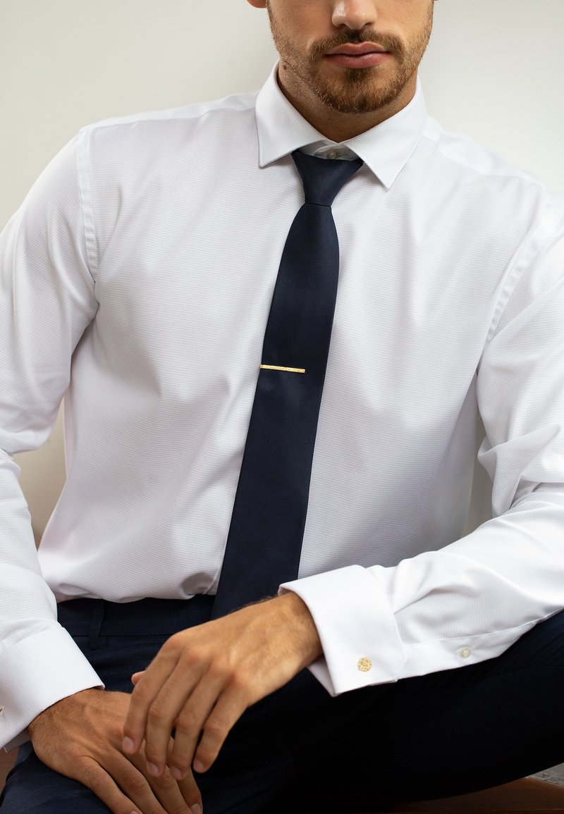 Weißes, strukturiertes Hemd mit einer navyblauen Krawatte und einer goldenen Krawattennadel; goldene Akzentuierung an der Manschette. Das Modell posiert mit einer entspannten Handhaltung.