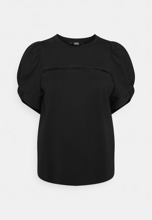 Sort bluse med korte ærmer med puffede ærmer og en vandret blonde detalje tværs over brystet mod en ensfarvet baggrund.
