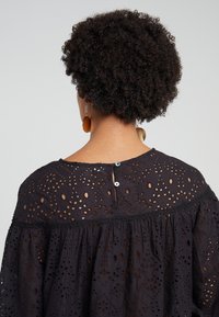 Blouse noire brodée avec des motifs découpés, des manches courtes et une fermeture à boutons au dos. Le tissu est léger et texturé.