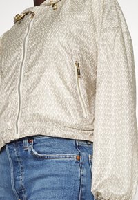 Giacca beige a fantasia con tasche frontali e laterali con zip dorate, indossata sopra jeans di denim blu.