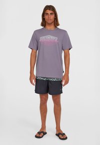 Lilla kortærmet t-shirt med et grafisk logo, parret med sorte badeshorts med et grønt logobånd, suppleret af sorte flip-flops.