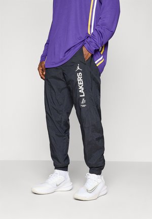 Homme portant un pantalon de survêtement noir des Los Angeles Lakers, un maillot de sport à manches longues violet, des baskets blanches, debout avec une main dans la poche.