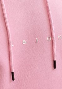 Hoodie rose pâle avec texte blanc indiquant "K & J" et cordons texturés. Présente une finition en tissu doux et un détail de couture subtil.