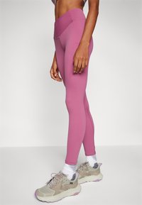 Hoch taillierte, enganliegende Leggings aus einem weichen pinkfarbenen Stoff, mit flachen Nähten und einer Seitentasche. Kombiniert mit hellgrauen Turnschuhen und weißen Socken.