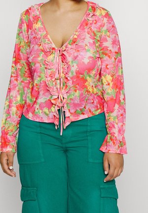 Woman wearing a long-sleeve pink floral ruffled blouse with ties, paired with high-waisted green cargo pants with front pockets.