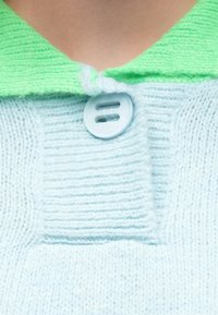 Light blue knit sweater with a green, textured collar, featuring a blue button. The fabric displays horizontal and vertical rib patterns.