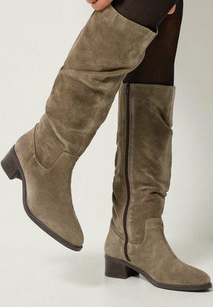 Bottes en daim taupe montant jusqu'au genou avec talons bas empilés et fermetures éclair latérales, portées sur des collants noirs, jambes croisées.