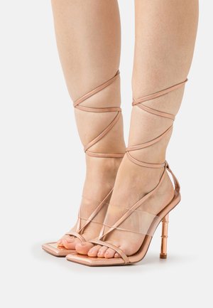 Sandales à talons hauts avec des brides, un devant transparent et des accents en or rose. orteils carrés et liens délicats autour de la cheville. Semelle texturée.
